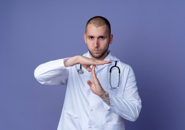 Médico varón joven disgustado vistiendo bata médica y estetoscopio alrededor de su cuello haciendo gesto de tiempo de espera aislado sobre fondo púrpura con espacio de copia