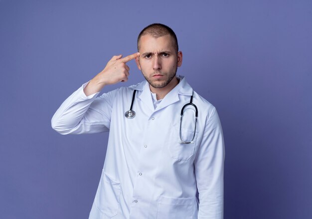 Médico varón joven disgustado vistiendo bata médica y estetoscopio alrededor de su cuello poniendo el dedo en la sien aislado sobre fondo púrpura con espacio de copia
