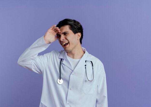 Médico varón joven disgustado vistiendo bata médica y un estetoscopio mirando al lado tocando la frente aislada