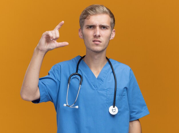 Médico varón joven disgustado vistiendo uniforme médico con estetoscopio mostrando tamaño aislado en pared naranja