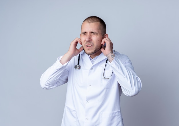 Foto gratuita médico varón joven molesto con bata médica y un estetoscopio alrededor de su cuello poniendo los dedos en las orejas con los ojos cerrados aislados en blanco con espacio de copia