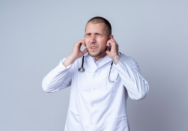 Médico varón joven molesto con bata médica y un estetoscopio alrededor de su cuello poniendo los dedos en las orejas con los ojos cerrados aislados en blanco con espacio de copia