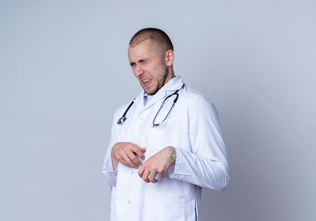 Médico varón joven molesto con bata médica y un estetoscopio alrededor de su cuello poniendo la mano sobre el vientre con los ojos cerrados aislados en blanco con espacio de copia