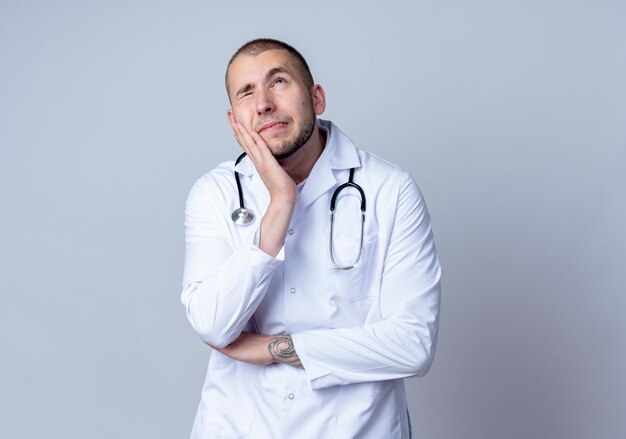 Médico varón joven pensativo con bata médica y estetoscopio alrededor de su cuello poniendo la mano en la mejilla mirando hacia arriba con un ojo cerrado aislado en la pared blanca