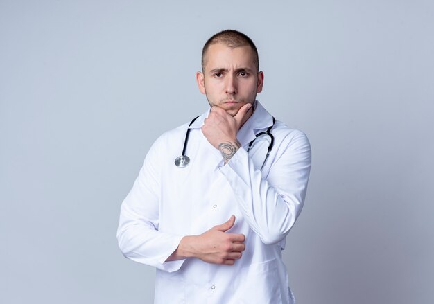 Médico varón joven pensativo con bata médica y un estetoscopio alrededor de su cuello poniendo la mano en el mentón aislado en la pared blanca