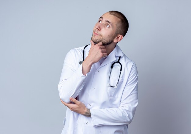 Médico varón joven pensativo con bata médica y un estetoscopio alrededor de su cuello tocando su barbilla y su codo mirando hacia arriba aislado en la pared blanca
