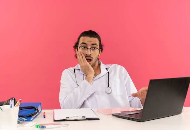Médico varón joven sorprendido con gafas médicas vistiendo bata médica con estetoscopio sentado en el escritorio