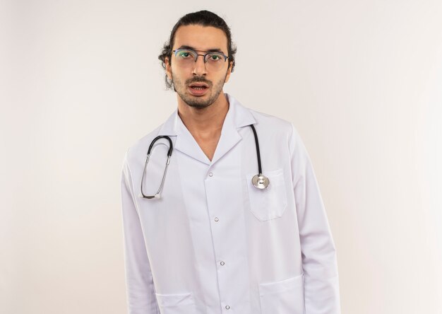Médico varón joven sorprendido con gafas ópticas con bata blanca con estetoscopio en pared blanca aislada con espacio de copia