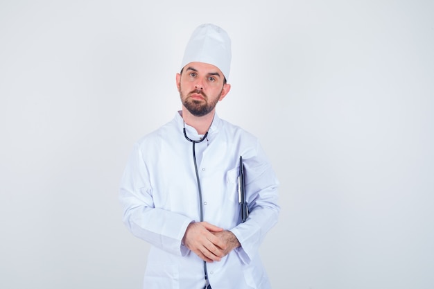 Médico varón joven sosteniendo el portapapeles en uniforme blanco y mirando serio, vista frontal.