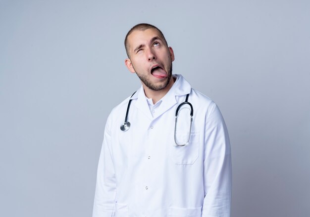 Médico varón joven tonto con bata médica y un estetoscopio alrededor de su cuello mirando hacia arriba y mostrando la lengua con un ojo cerrado aislado en la pared blanca