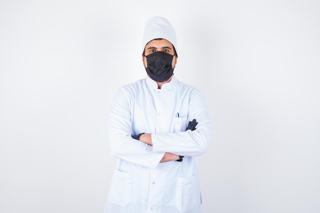 Médico varón joven en uniforme blanco de pie con los brazos cruzados y mirando confiado, vista frontal.