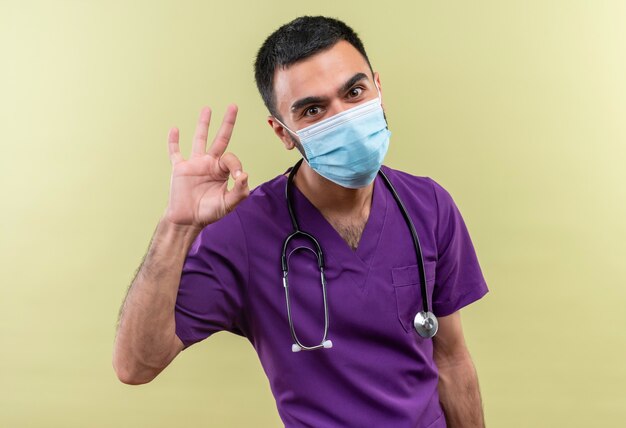 Médico varón joven vistiendo ropa de cirujano púrpura y estetoscopio máscara médica mostrando gesto okey en la pared verde aislada