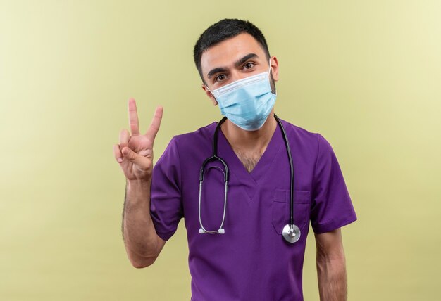 Médico varón joven vistiendo ropa de cirujano púrpura y estetoscopio máscara médica mostrando gesto de paz en la pared verde aislada