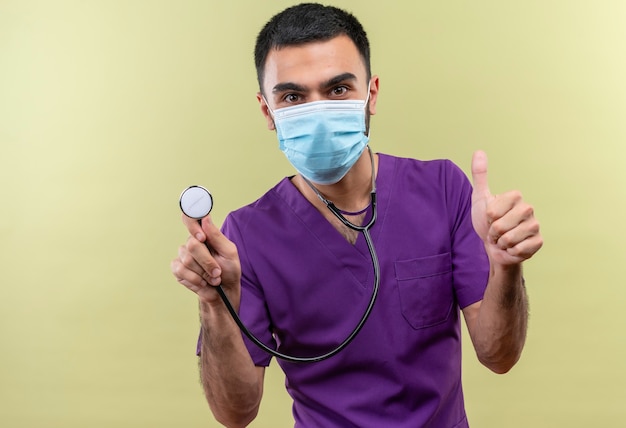 Médico varón joven vistiendo ropa de cirujano púrpura y estetoscopio máscara médica con el pulgar hacia arriba en la pared verde aislada