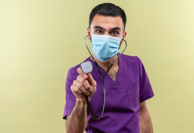 Médico varón joven vistiendo ropa de cirujano púrpura y estetoscopio máscara médica sosteniendo estetoscopio en la pared verde aislada