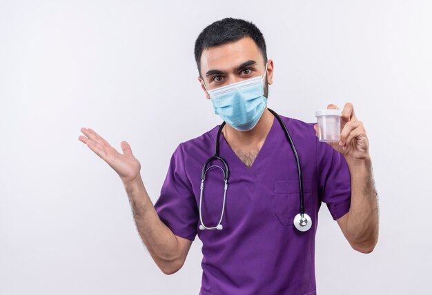 Médico varón joven vistiendo ropa de cirujano púrpura y estetoscopio máscara médica sosteniendo lata vacía en la pared blanca aislada