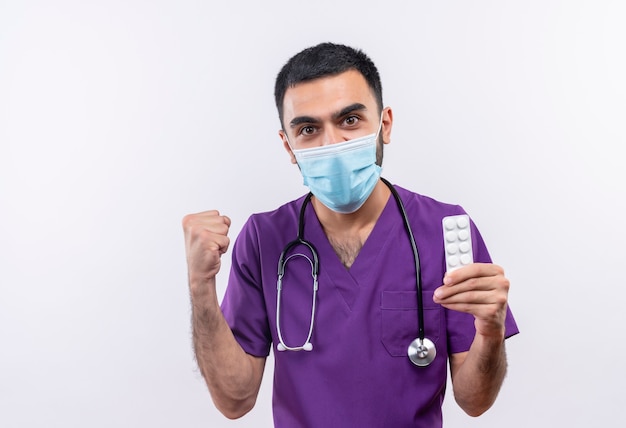 Médico varón joven vistiendo ropa de cirujano púrpura y estetoscopio máscara médica sosteniendo pastillas que muestran gesto de sí en la pared blanca aislada