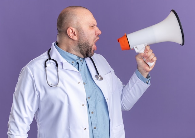 Médico varón de mediana edad con bata médica y un estetoscopio mirando al lado gritando en altavoz aislado en la pared púrpura