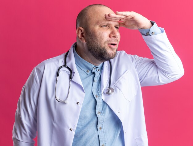 Médico varón de mediana edad concentrado con bata médica y un estetoscopio manteniendo la mano en la frente mirando al lado en la distancia aislada en la pared rosa