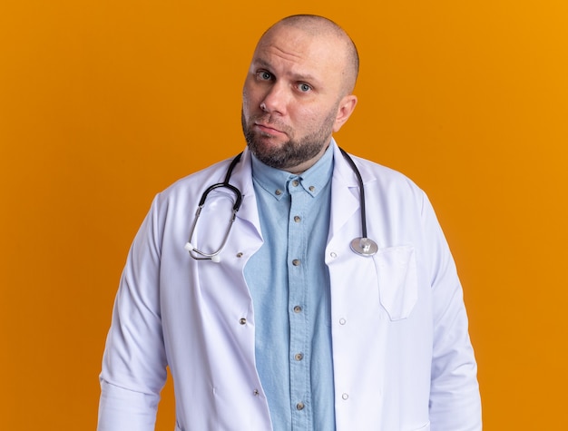 Médico varón de mediana edad confundido con bata médica y estetoscopio aislado en la pared naranja
