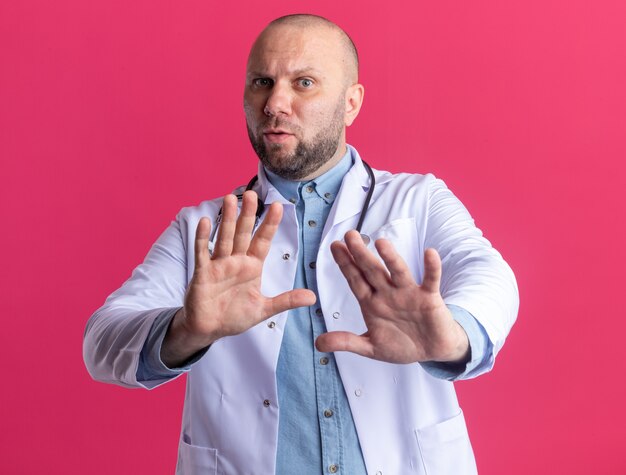 Médico varón de mediana edad disgustado con bata médica y estetoscopio haciendo gesto de rechazo aislado en la pared rosa