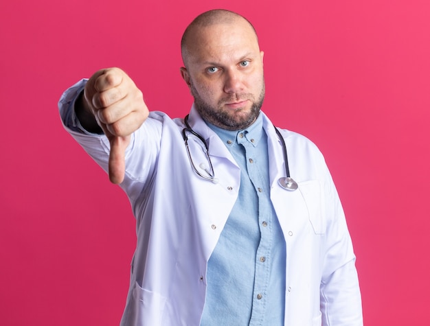 Médico varón de mediana edad disgustado con bata médica y un estetoscopio mirando al frente mostrando el pulgar hacia abajo aislado en la pared rosa