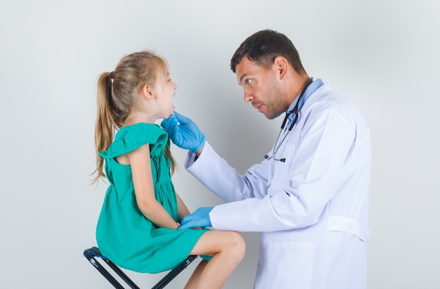 Médico varón mirando en la boca de la niña en uniforme blanco, guantes