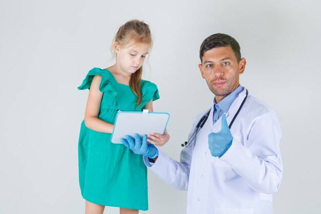 Médico varón mostrando el pulgar hacia arriba mientras el niño escribe a bordo en bata blanca