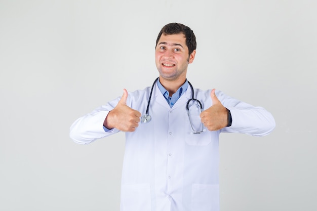 Médico varón mostrando los pulgares para arriba en bata blanca y mirando alegre