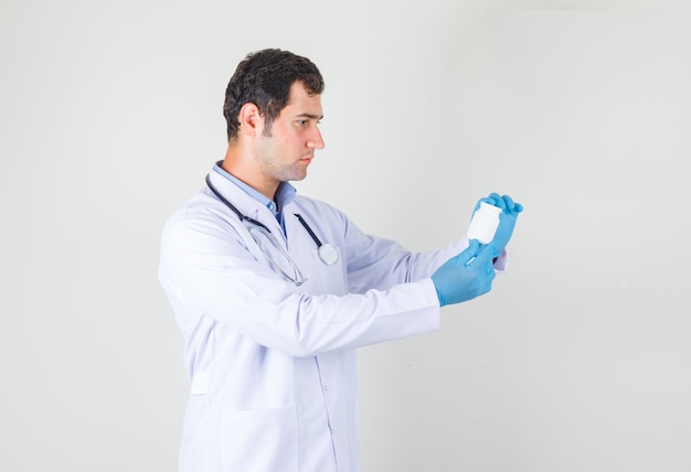 Médico varón sosteniendo una botella de píldoras en bata blanca, guantes y mirando serio