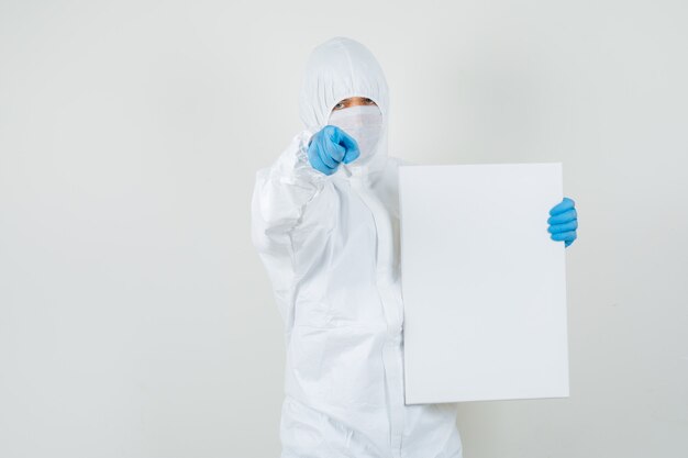 Médico varón sosteniendo un lienzo en blanco, apuntando a la cámara en traje protector