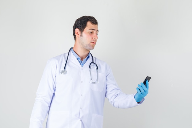 Médico varón sosteniendo smartphone en bata blanca, guantes y mirando serio