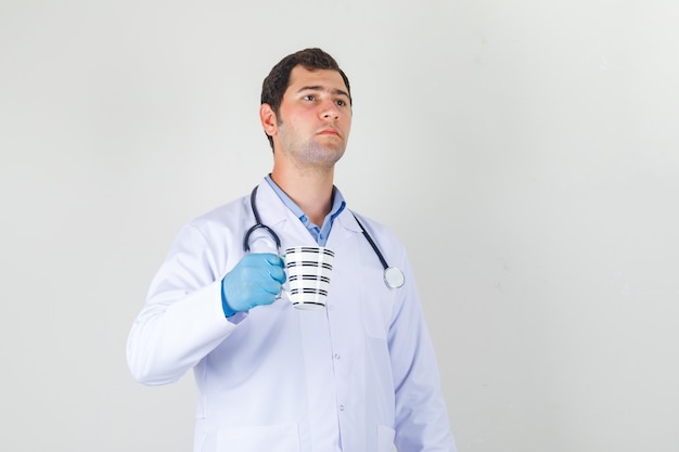 Médico varón sosteniendo una taza de bebida en bata blanca, guantes y mirando pensativo
