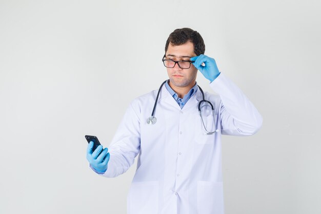 Médico varón sosteniendo el teléfono inteligente con la mano en las gafas en bata blanca, guantes