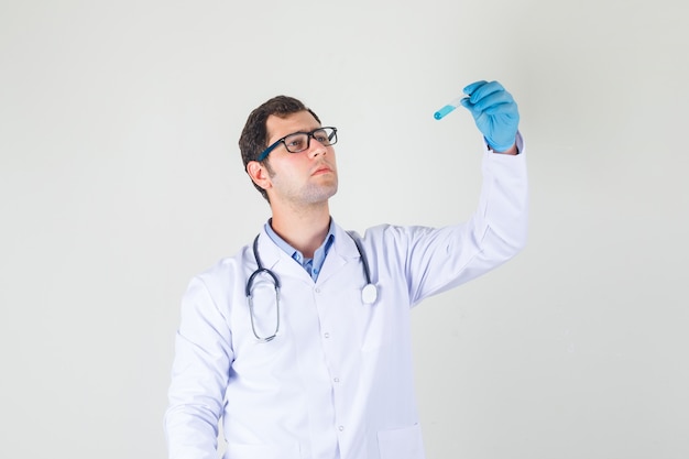 Médico varón sosteniendo el tubo de ensayo en bata blanca, guantes, gafas y mirando serio