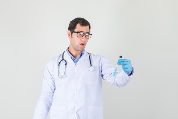 Médico varón sosteniendo el tubo de ensayo en bata blanca, guantes, gafas y mirando sorprendido. vista frontal.