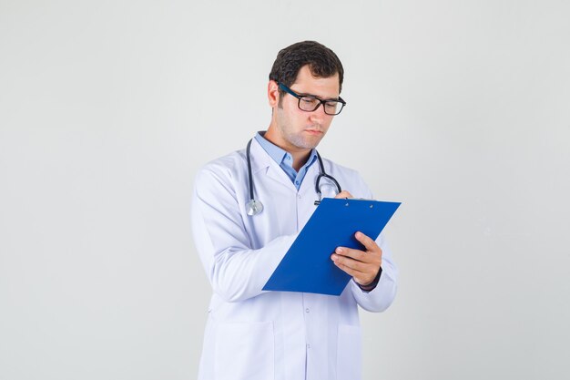 Médico varón tomando notas en el portapapeles en bata blanca, gafas y mirando ocupado. vista frontal.