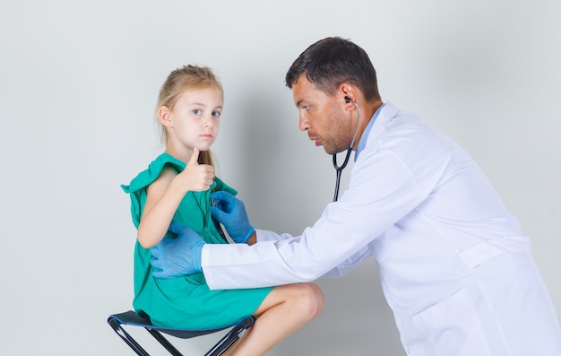 Médico varón en uniforme blanco escuchando latidos mientras el niño mostrando el pulgar hacia arriba