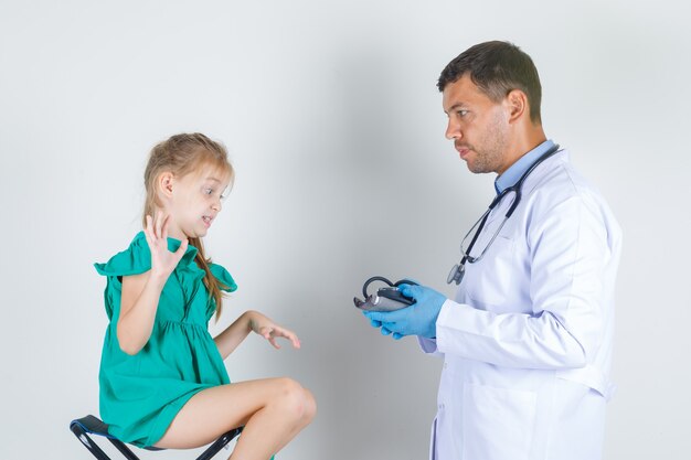 Médico varón en uniforme blanco, guantes sosteniendo esfigmomanómetro mientras el niño mira con disgusto