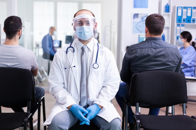 Médico con visera contra coronavirus en sala de espera mirando cámara de introducción