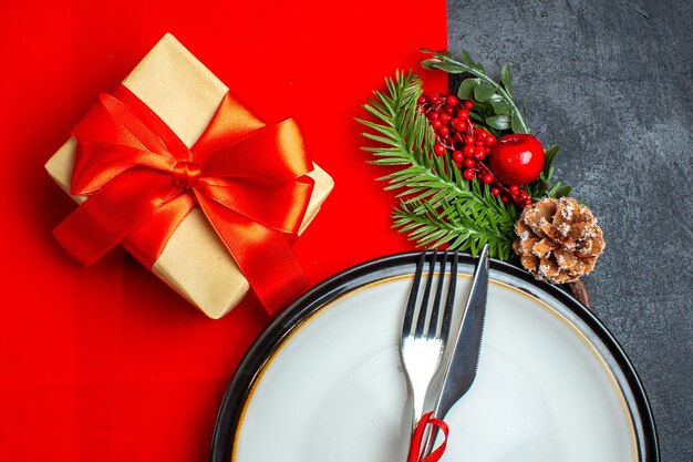 Medio plano de fondo de año nuevo con cubiertos con cinta roja en un plato de cena accesorios de decoración ramas de abeto junto a un regalo en una servilleta roja