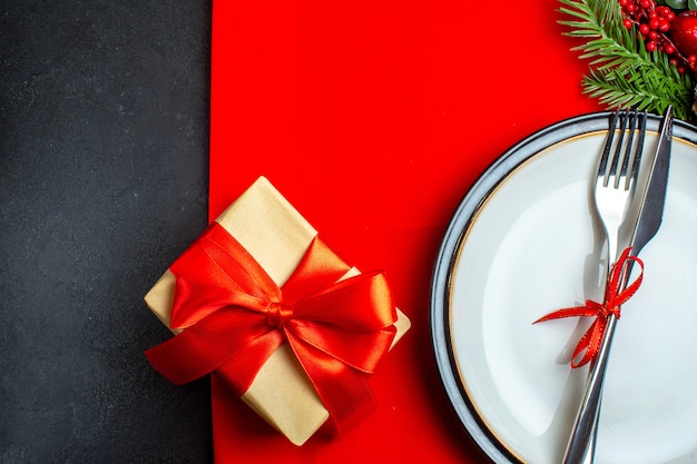 Medio plano de fondo de Navidad con cubiertos con cinta roja sobre un plato de cena accesorios de decoración ramas de abeto junto a un regalo en una servilleta roja