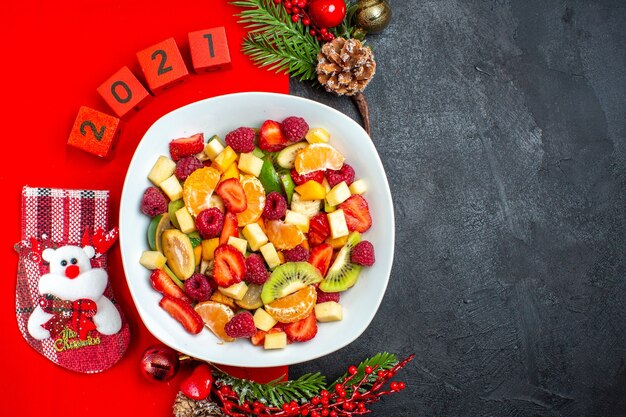 Medio tiro de la colección de frutas frescas en accesorios de decoración de plato de cena ramas de abeto y números en una servilleta roja en el lado derecho sobre fondo oscuro