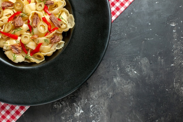 Medio tiro de deliciosa conchiglie con verduras en un plato negro y un cuchillo sobre una toalla despojada de rojo sobre la mesa gris