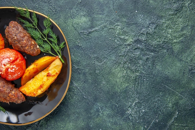 Medio tiro de deliciosas chuletas de carne al horno con patatas y tomates en una placa negra en el lado derecho sobre fondo de color verde negro mezclado