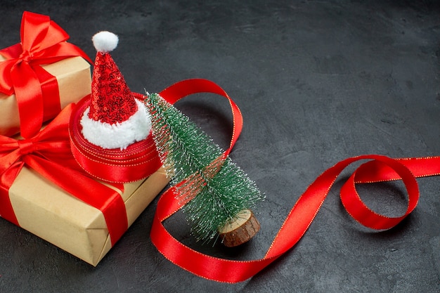 Medio tiro de hermosos regalos con cinta roja y sombrero de santa claus árbol de navidad en mesa oscura