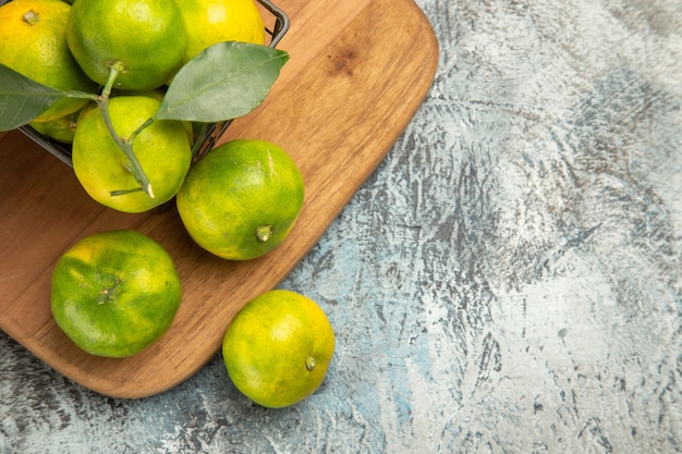 Medio tiro de mandarinas verdes con hojas dentro y fuera de una canasta en la tabla de cortar de madera en la mesa gris