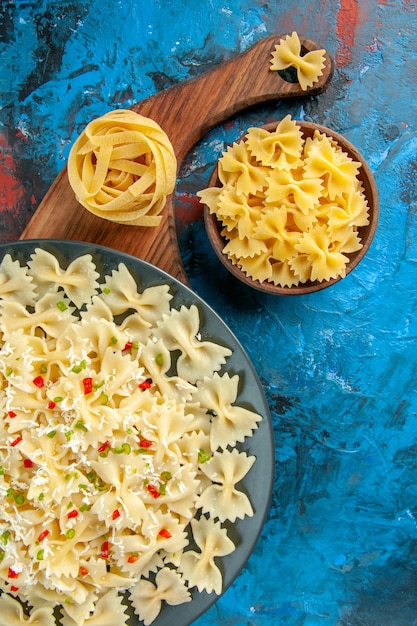 Medio tiro de pastas farfalle con verduras en una placa negra sobre tabla de cortar de madera y varias pastas sobre fondo azul.