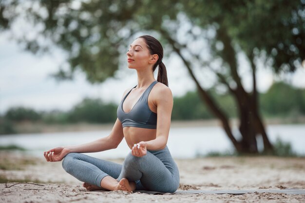 Meditando con sonidos de la naturaleza. Morena con forma de cuerpo agradable en ropa deportiva tiene día de fitness en la playa