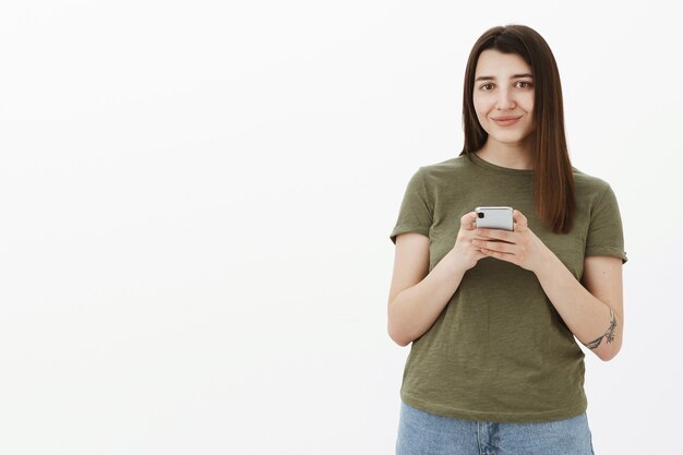 La mejor aplicación de todas. Retrato de mujer joven agradable contenta y optimista con cabello castaño en camiseta verde oliva sonriendo satisfecha como sosteniendo un teléfono inteligente con expresión encantada, usando un nuevo dispositivo